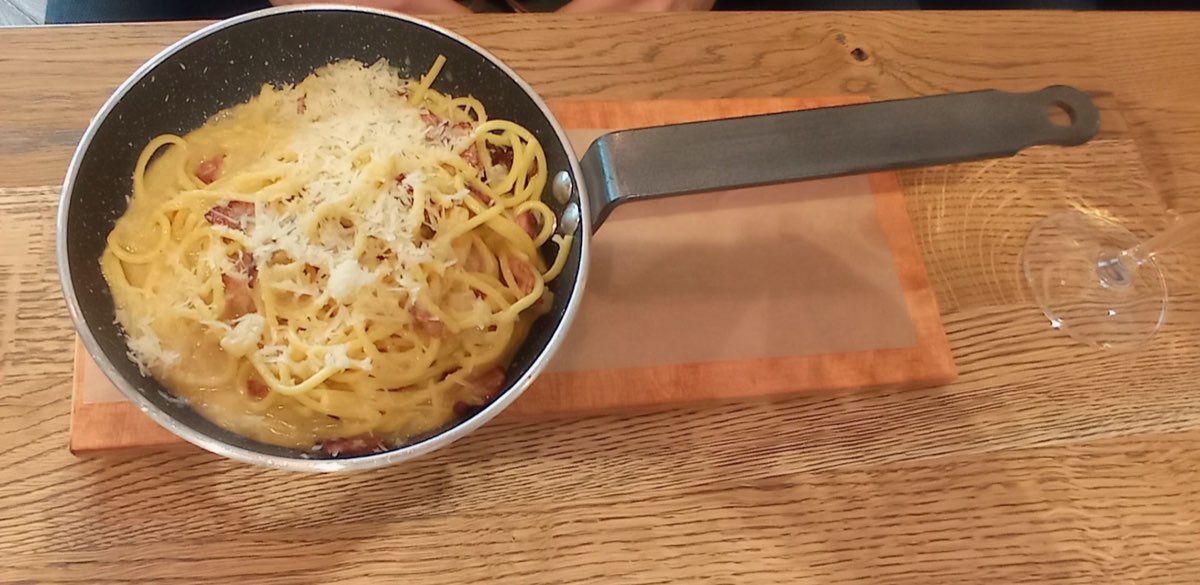 Spaghetti carbonara z parmezanem, podane prosto na patelni na drewnianej desce.